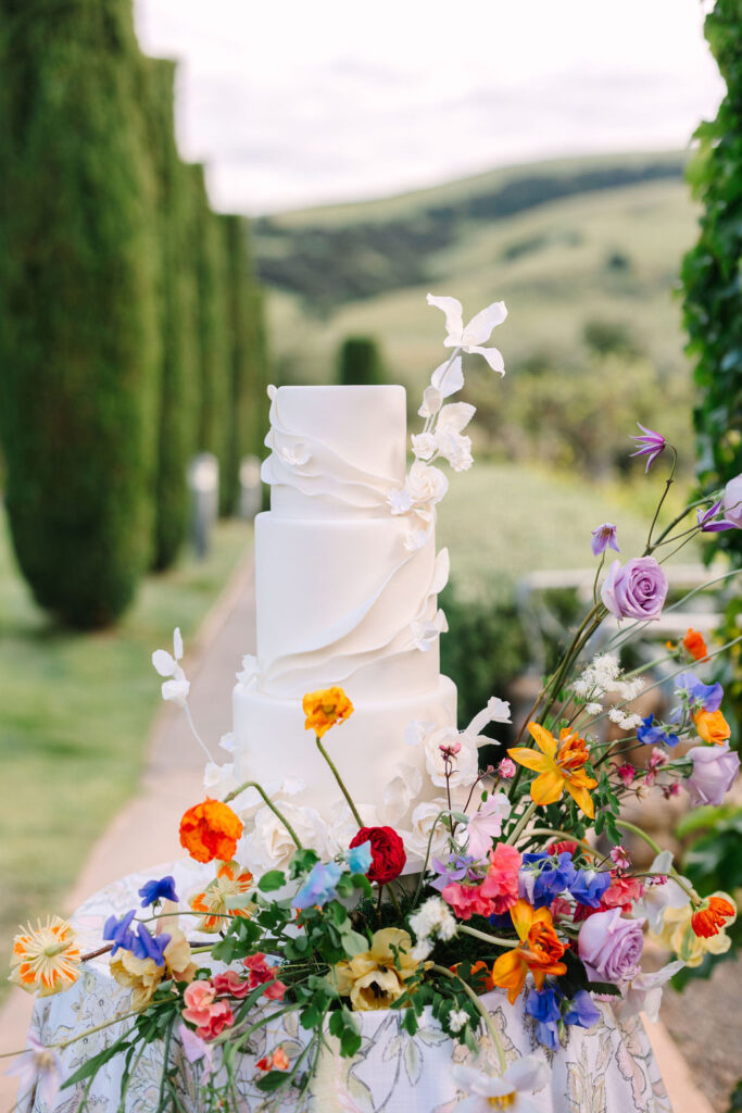 A three-tiered white wedding cake with floral decorations stands on a table with a colorful flower arrangement. Trees and rolling hills are in the background.