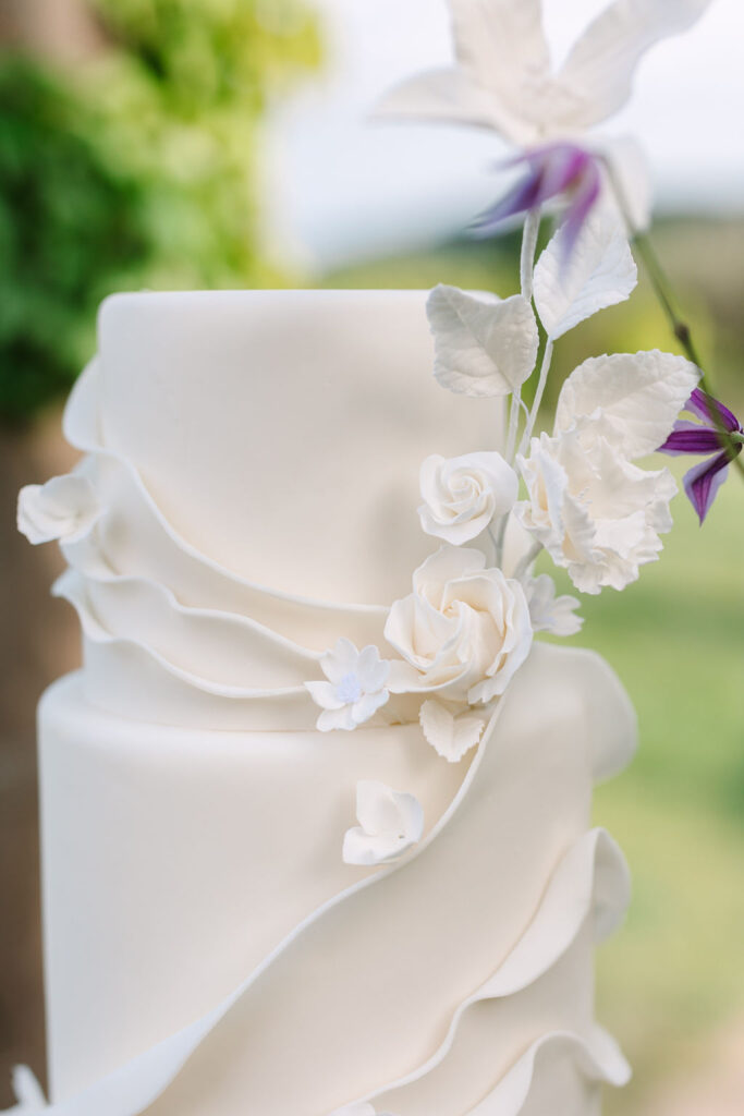 Three-tier white cake with delicate white floral decorations and cascading fondant ruffles, set outdoors.