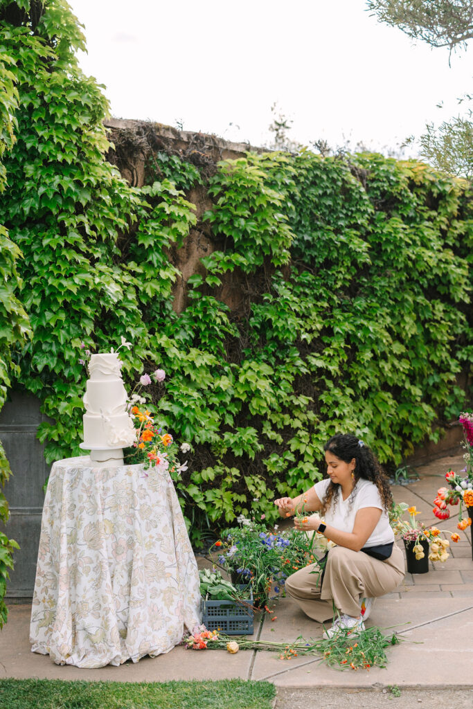 A woman arranges flowers beside a decorated table with a three-tiered white cake, set against a backdrop of greenery.