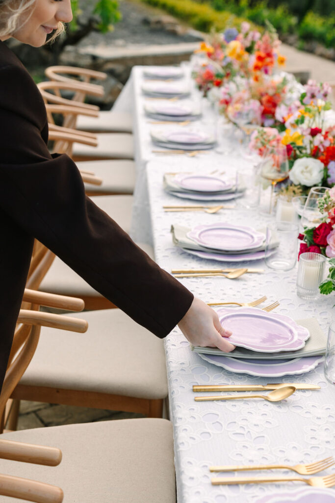 A person sets a lavender plate on an outdoor dining table adorned with a white lace cloth, gold cutlery, and colorful floral arrangements.