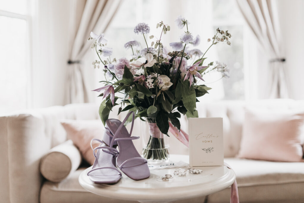 Purple sandals, a bouquet of pink and white flowers, a "Vows" booklet, and jewelry are arranged on a small round table in front of a sofa and large windows.