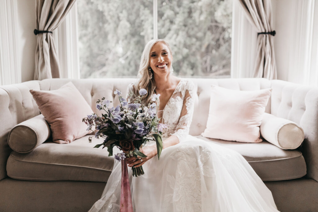A woman in a white wedding dress sits on a sofa holding a bouquet of purple flowers. Beige curtains frame a window in the background.