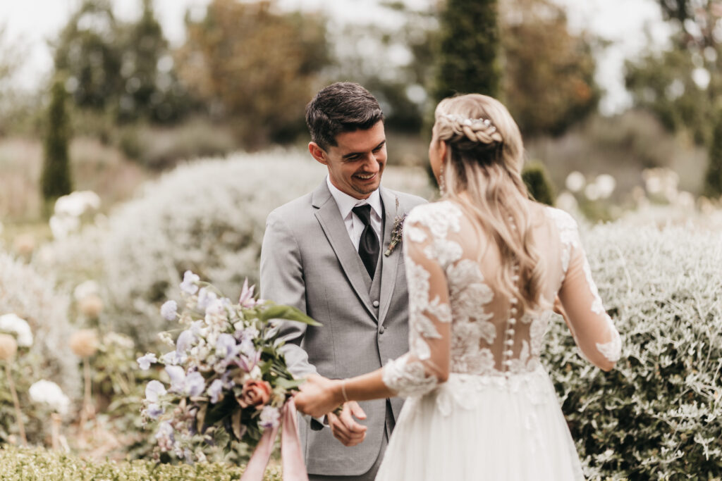 A bride and groom stand facing each other outdoors. The groom, in a gray suit, smiles at the bride, who holds a bouquet and wears a dress with lace detailing. They're surrounded by greenery.