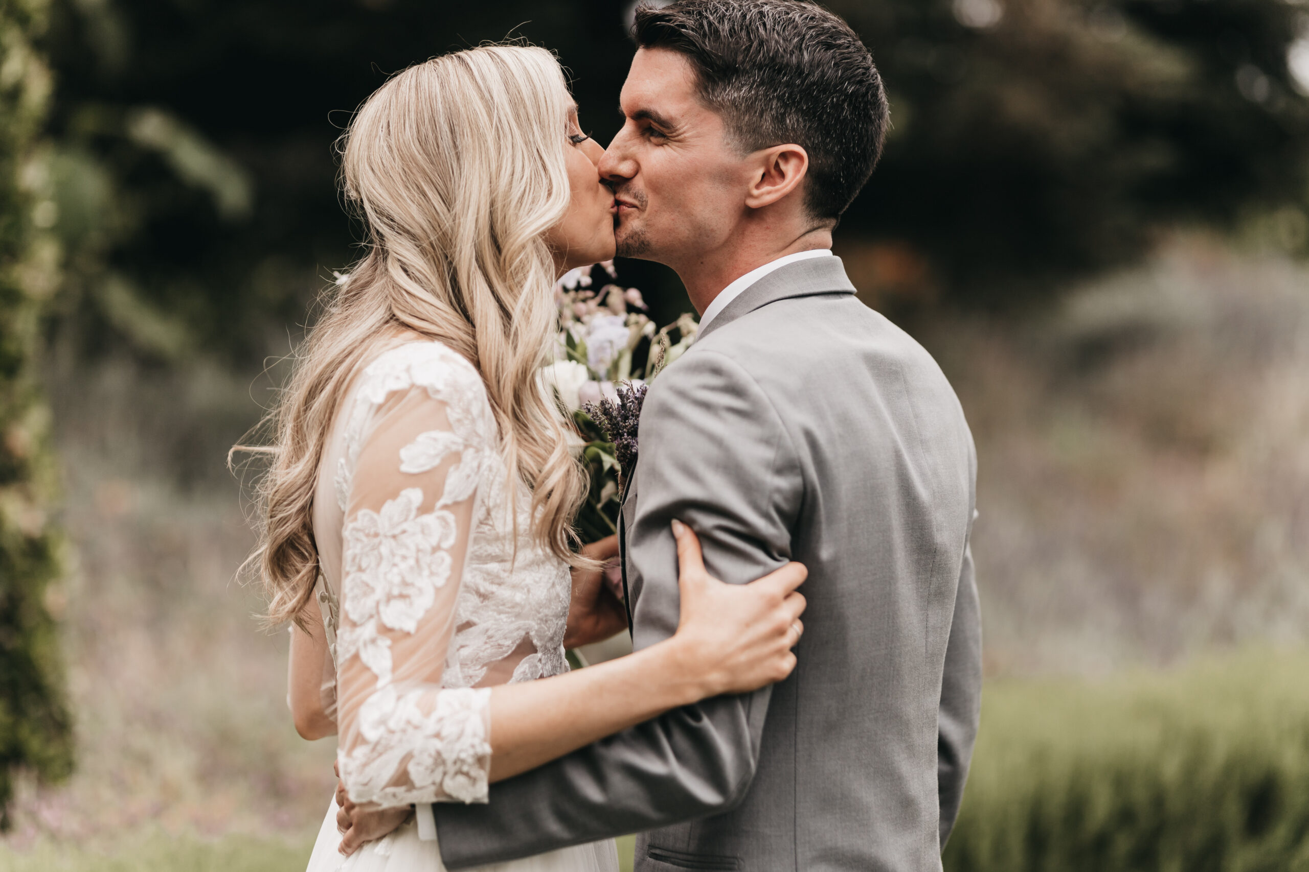 A couple, dressed formally, shares a kiss outdoors. The woman holds a bouquet, and both have closed eyes.