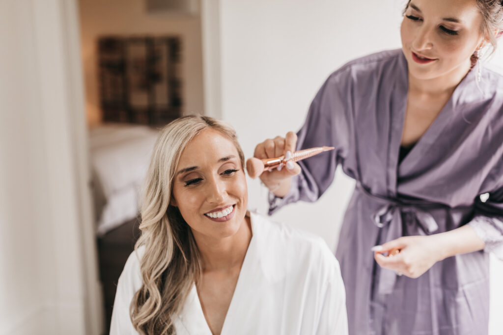 A woman in a white robe smiles while another woman in a purple robe styles her hair with a curling iron.