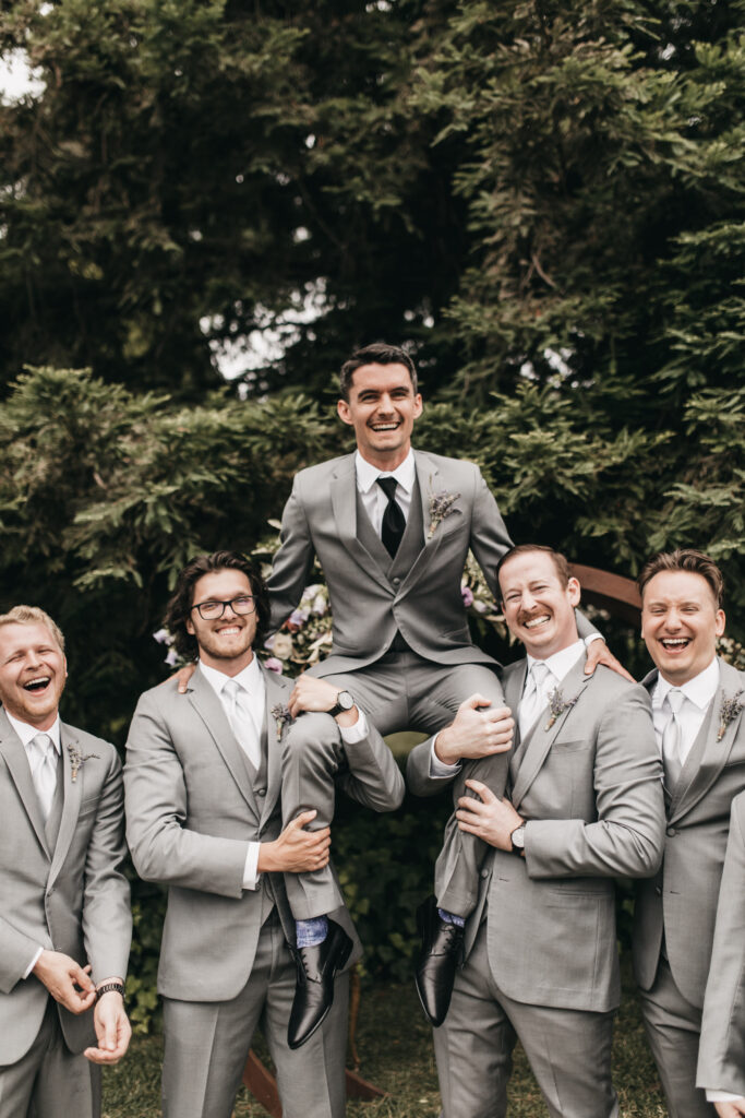 Five men in matching grey suits, one being lifted by the others, pose cheerfully in front of lush greenery.