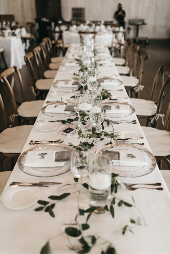 A long dining table set for an event, with white tablecloth, elegant plates, glasses, cutlery, and green foliage running down the center. Wooden chairs surround the table.