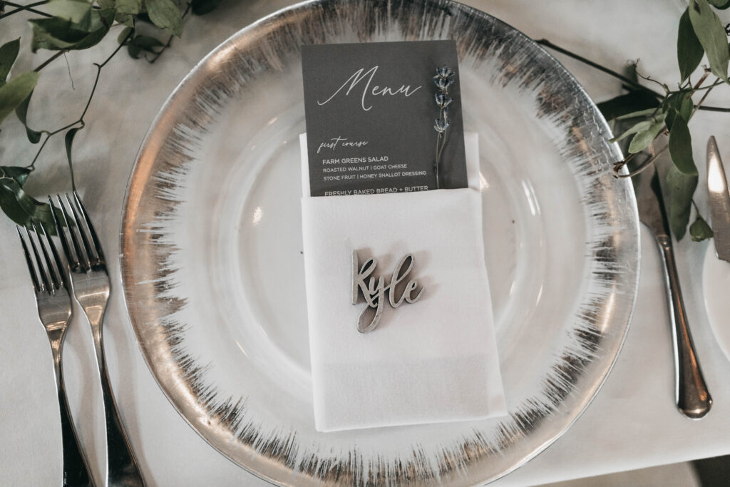 Place setting with a metallic-rimmed plate, personalized name card "Kyle," menu, and stainless steel cutlery, set on a white tablecloth with green leaves.