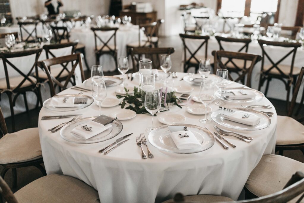 A round table set for a formal event with white tablecloth, neatly arranged plates, glassware, cutlery, and small place cards, surrounded by wooden chairs.