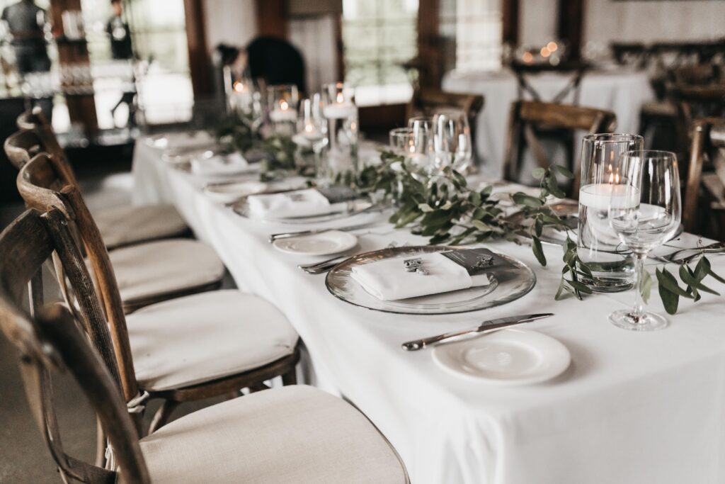 A long dining table with white tablecloth, set with plates, silverware, glasses, and candles. Greenery decorates the center. Wooden chairs are arranged around the table.