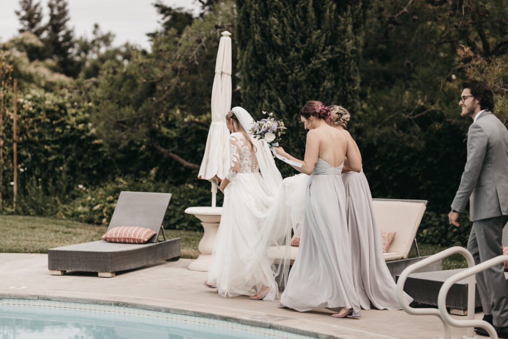 Bride in white gown and veil walks by a pool with bridesmaids in lavender dresses and a man in a gray suit, set against a lush garden backdrop.