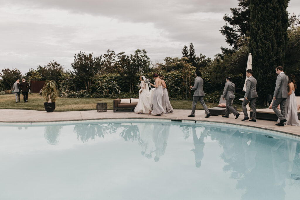 A wedding party in formal attire walks by a poolside, surrounded by greenery under an overcast sky.