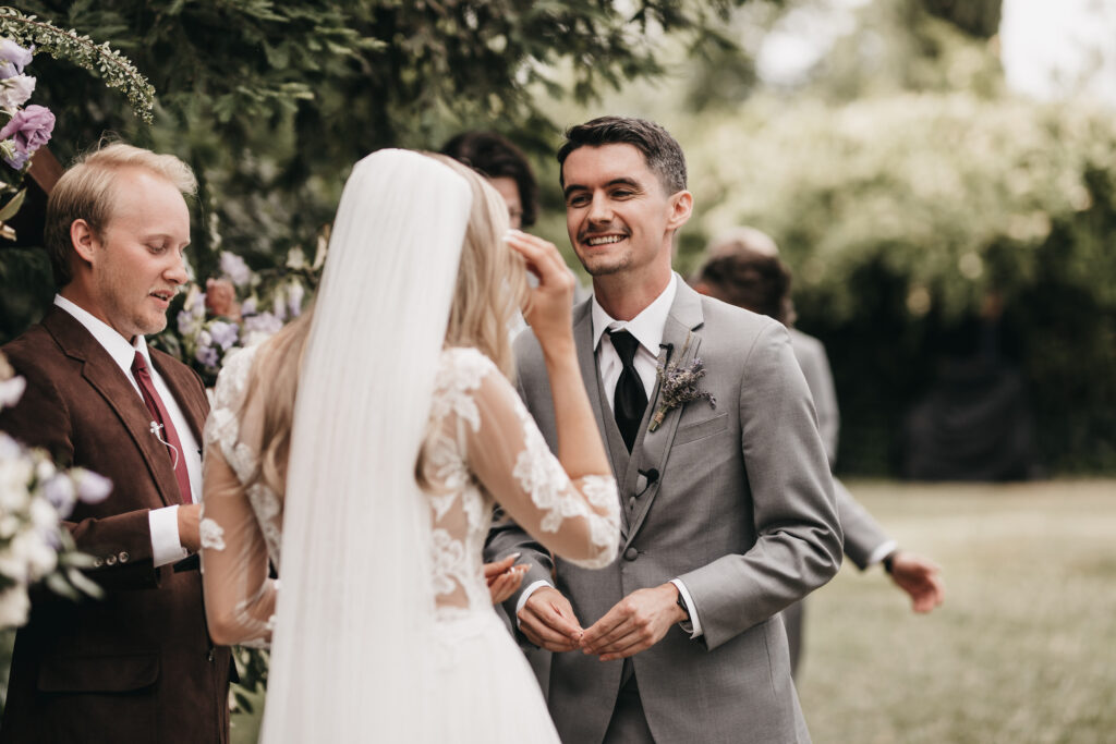 A bride and groom stand together outdoors. The groom, in a gray suit, is smiling. The bride, in a white dress and veil, is facing him. Another person in a brown suit is nearby.