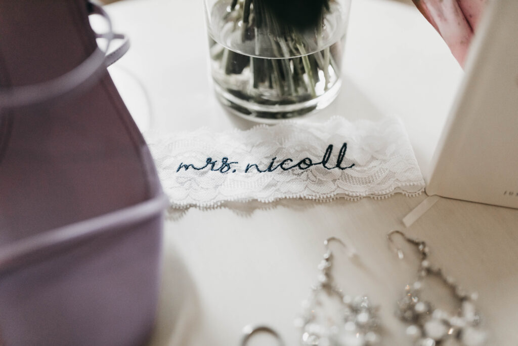 Lace fabric with "mrs. nicoll" embroidered on it, placed on a white surface near a purple box, glass vase, and silver earrings.
