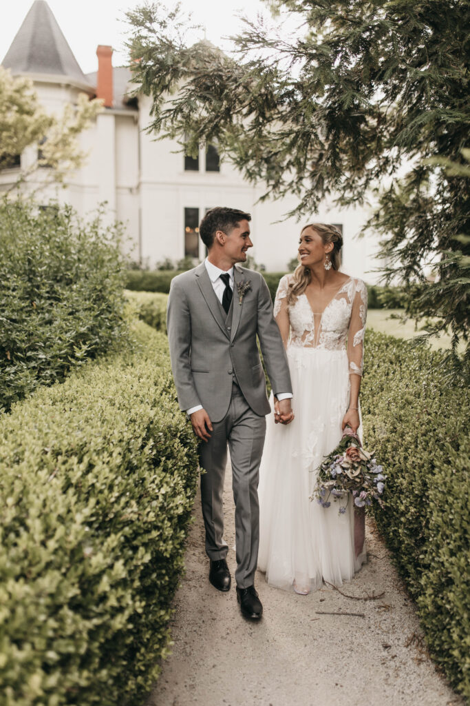 A bride and groom walk hand in hand along a garden path. The groom is in a gray suit, and the bride wears a white lace dress, holding a bouquet. They are surrounded by greenery.