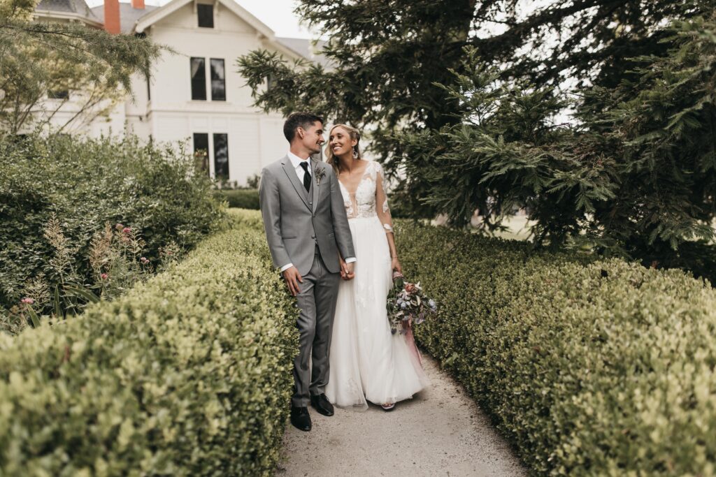 A couple in wedding attire holds hands while walking down a path surrounded by greenery and a white house in the background.
