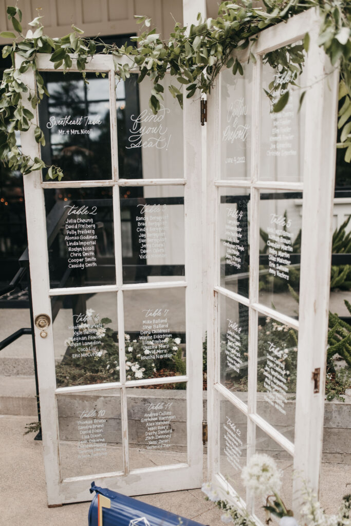 Vintage doors are used as a wedding seating chart, with guest names written on the glass panes. Greenery decorates the top.