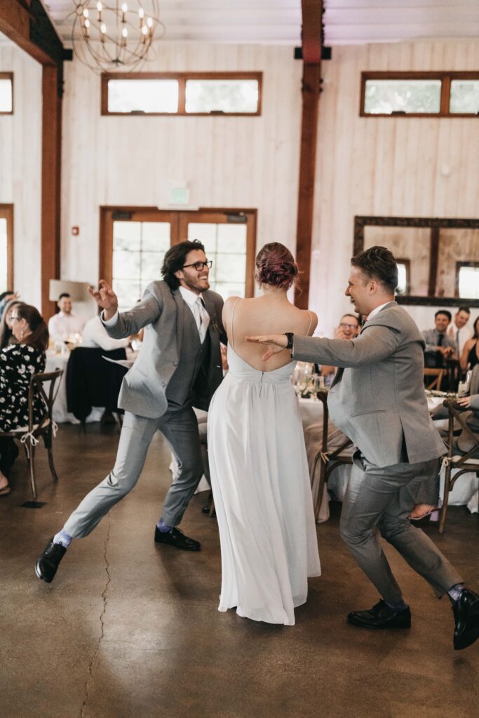 Three people in formal attire dance with enthusiasm in a bright indoor venue, surrounded by seated guests.