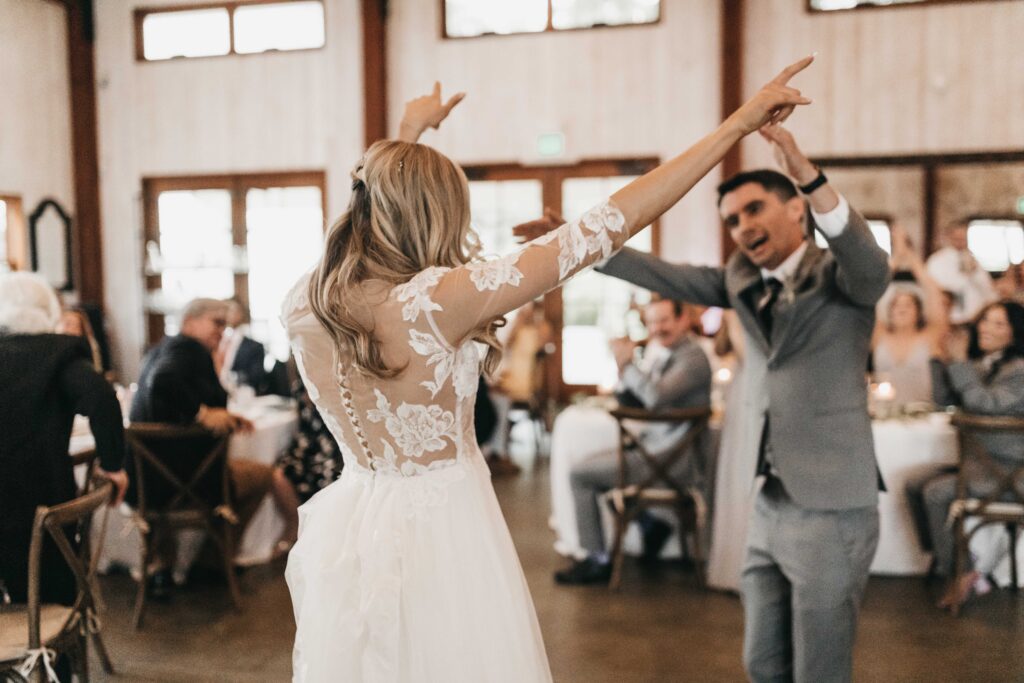 A bride in a white lace dress and a groom in a gray suit dance joyfully, with guests seated at tables in the background.