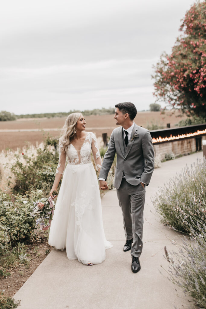 A bride in a white gown and a groom in a gray suit walk hand in hand down a path lined with plants, with fields and a floral backdrop.