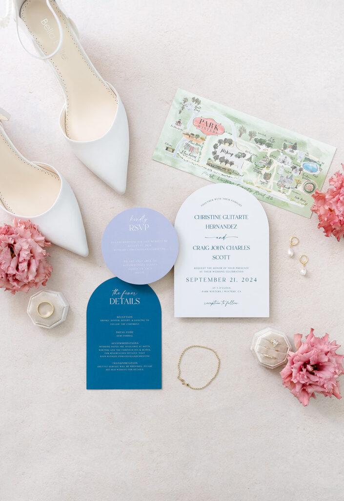Flat lay of wedding invitation suite with RSVP card, blue details card, map, white heels, pink flowers, earrings, bracelet, and a ring on a light surface.
