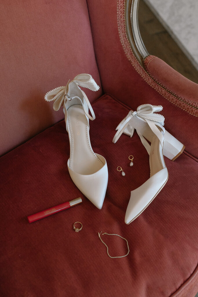 White high-heeled shoes with ribbon details, jewelry, and a red lip product are arranged on a pink upholstered chair.