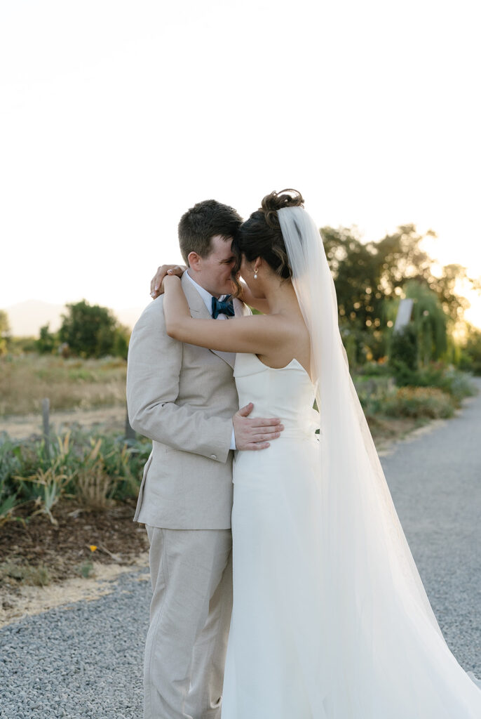 A bride and groom embrace outdoors on a gravel path, dressed in a white gown and beige suit, with greenery and trees in the background.