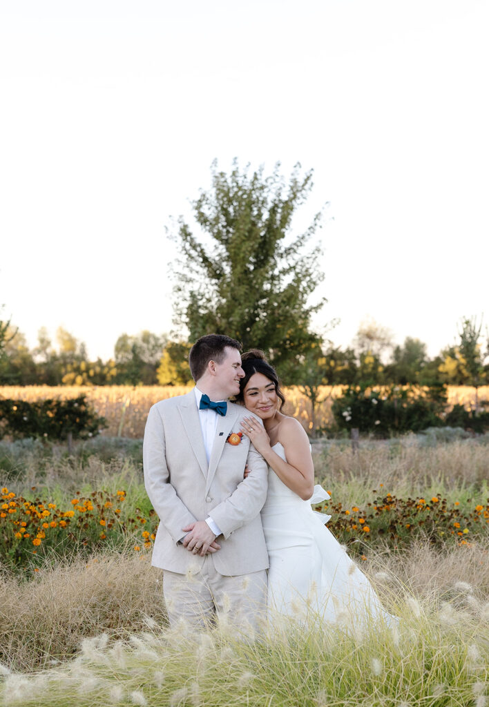 A bride in a white dress leans on a groom in a beige suit as they stand in a field with wildflowers and tall grass, with trees and clear sky in the background.