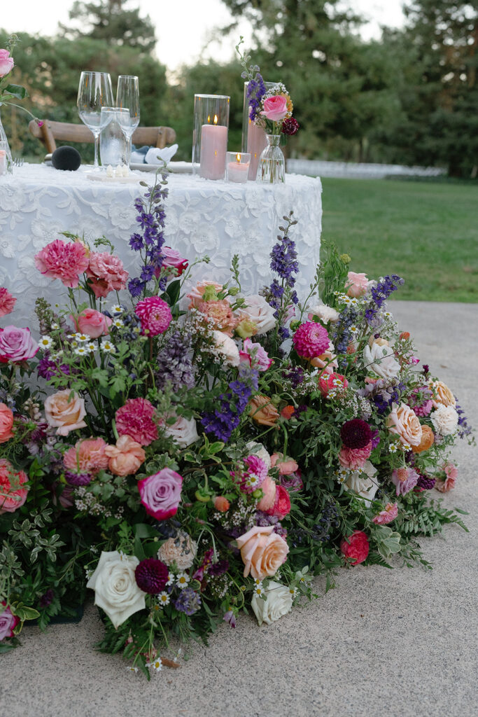 A white table with glassware and candles is decorated with an arrangement of pink, purple, and white flowers and greenery outdoors.