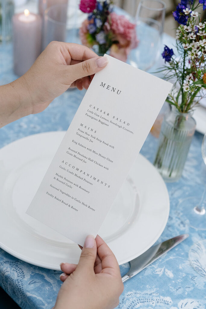 A person holds a printed wedding menu above a white plate on a set table with blue lace tablecloth, candles, and vases of flowers.
