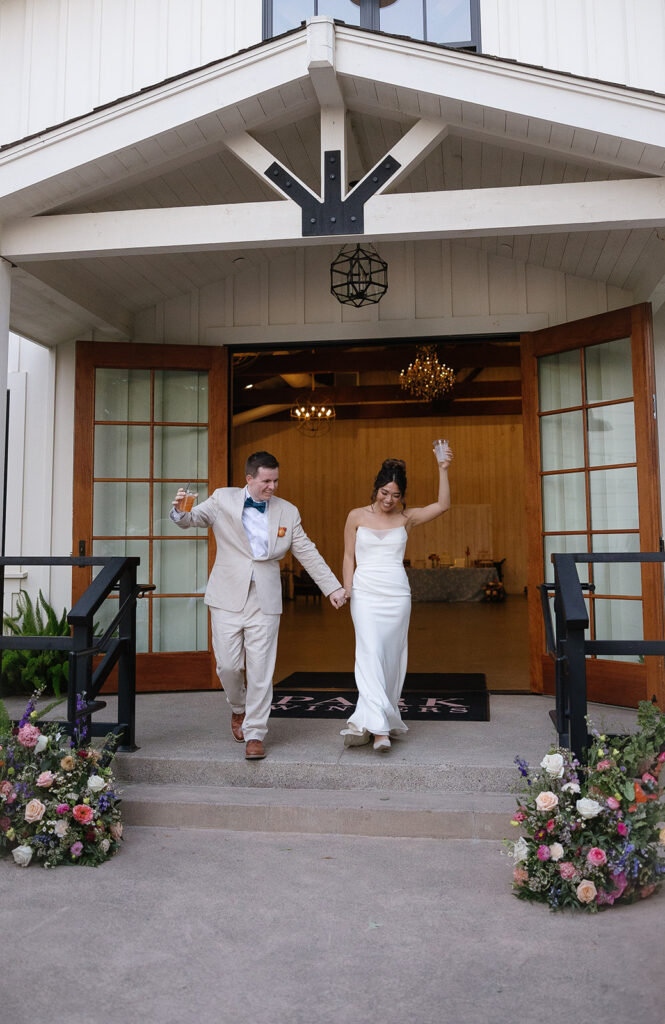 A couple in formal attire exits a building holding hands; the woman waves, and the man holds a drink. Floral arrangements frame the entrance.