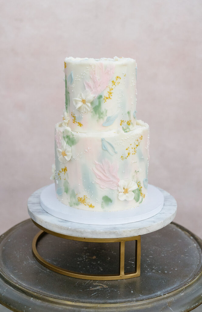 Two-tiered cake with white icing, pastel floral decorations, small white flowers, and gold accents, displayed on a marble and metal cake stand.