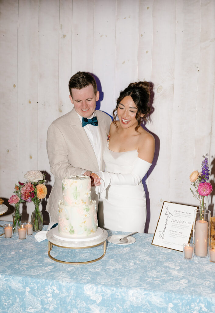 A bride and groom stand together, smiling as they cut a two-tiered wedding cake on a decorated table with candles, flowers, and a dessert menu.