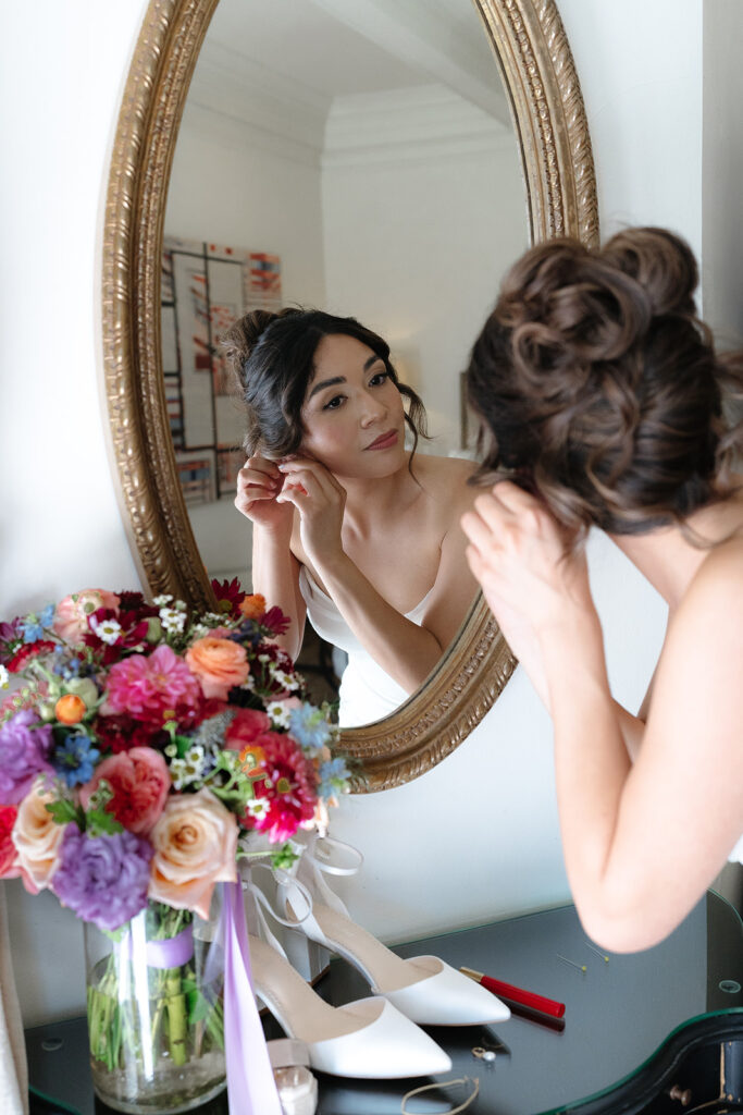 A woman in a white dress adjusts her earring while looking in a mirror. A bouquet of colorful flowers and a pair of white high-heeled shoes are on the table in front of her.