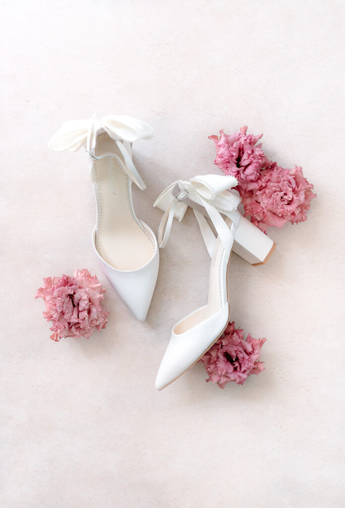 A pair of white high-heeled shoes with ankle bows, arranged among four pink ruffled flowers on a light background.