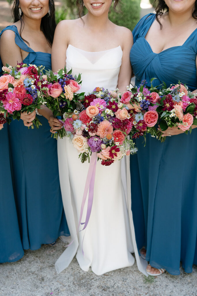 Three women in blue and white dresses hold colorful floral bouquets, standing outdoors. The focus is on their dresses and flowers, not their faces.