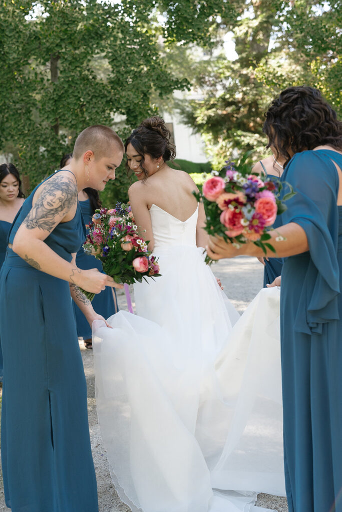 A bride in a white dress is assisted by bridesmaids in teal dresses, holding bouquets and adjusting her train outdoors among trees.