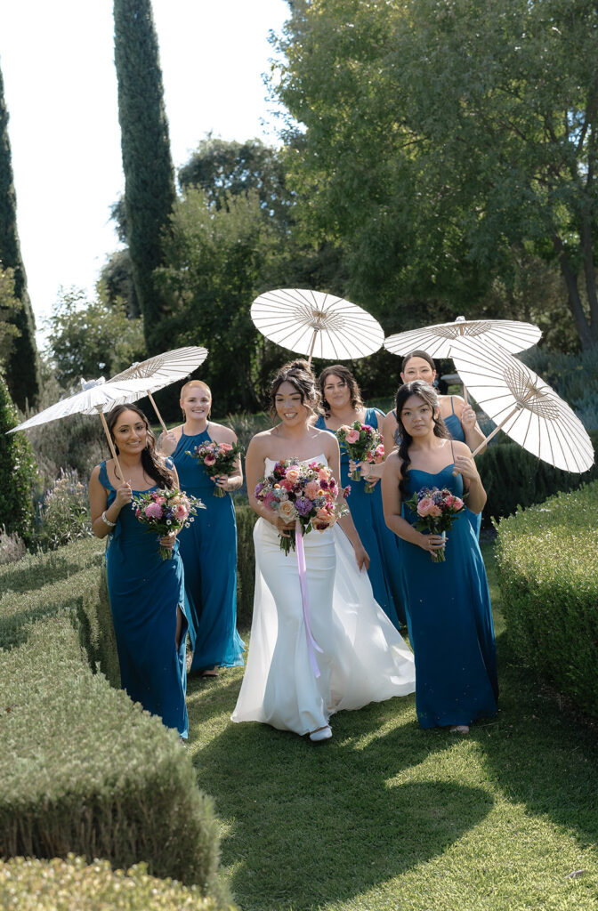 A bride in a white dress walks outside with five bridesmaids in teal dresses holding parasols and bouquets, surrounded by greenery on a sunny day.
