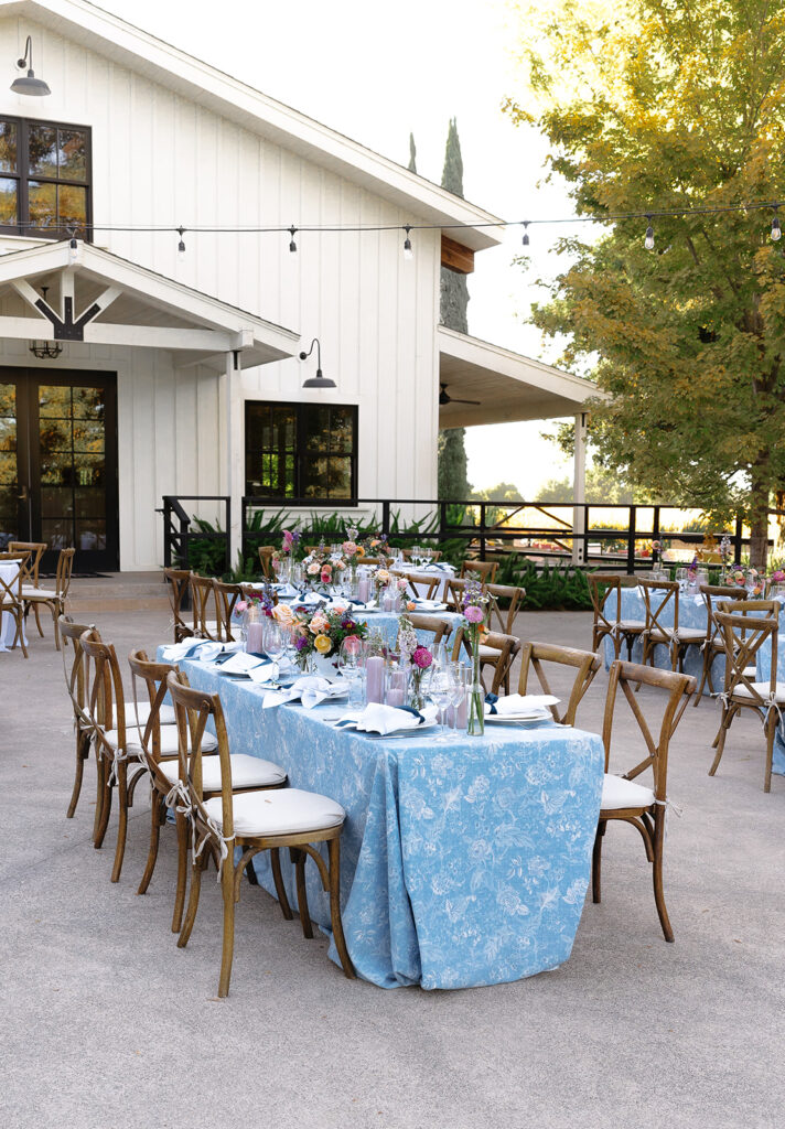 Rectangular outdoor table set with blue-patterned cloth, floral centerpieces, and wooden chairs arranged beside a modern white building.