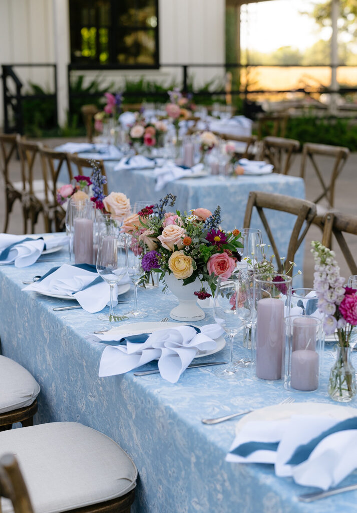 Outdoor table set for an event with blue tablecloths, floral centerpieces, napkins, glassware, and purple candles; wooden chairs surround the tables.