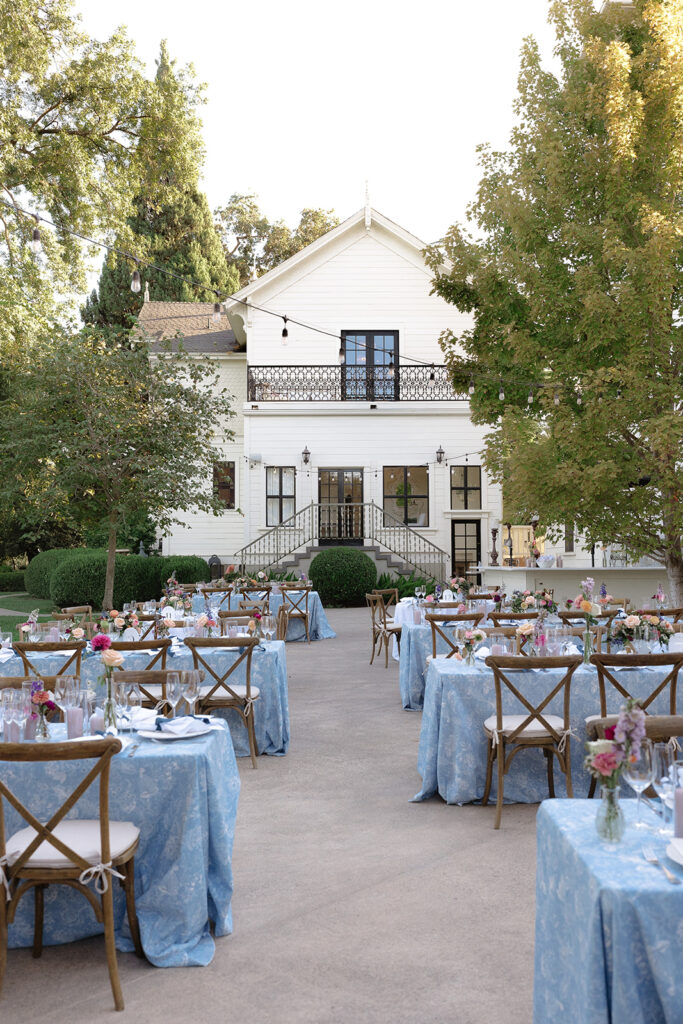 Round and square tables with blue tablecloths and floral centerpieces are arranged outdoors in front of a two-story white house with stairs and a balcony.