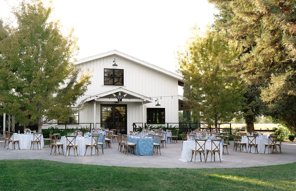 Outdoor wedding reception setup with round tables covered in white and blue linens, wooden chairs, and place settings in front of a white barn surrounded by trees.