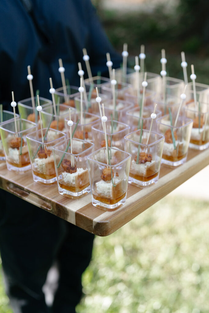 A person holds a wooden tray with multiple small clear cups containing layered appetizers, each with a skewer.