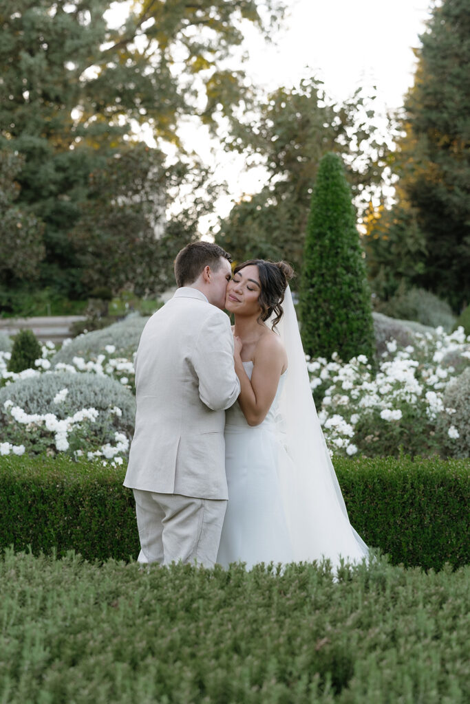 A bride and groom embrace and share a kiss in a garden with white flowers and greenery in the background.