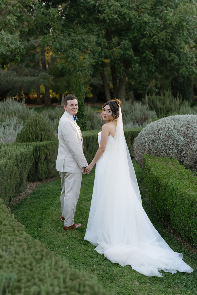 A bride and groom hold hands and smile while standing on a garden path, surrounded by green hedges and trees.