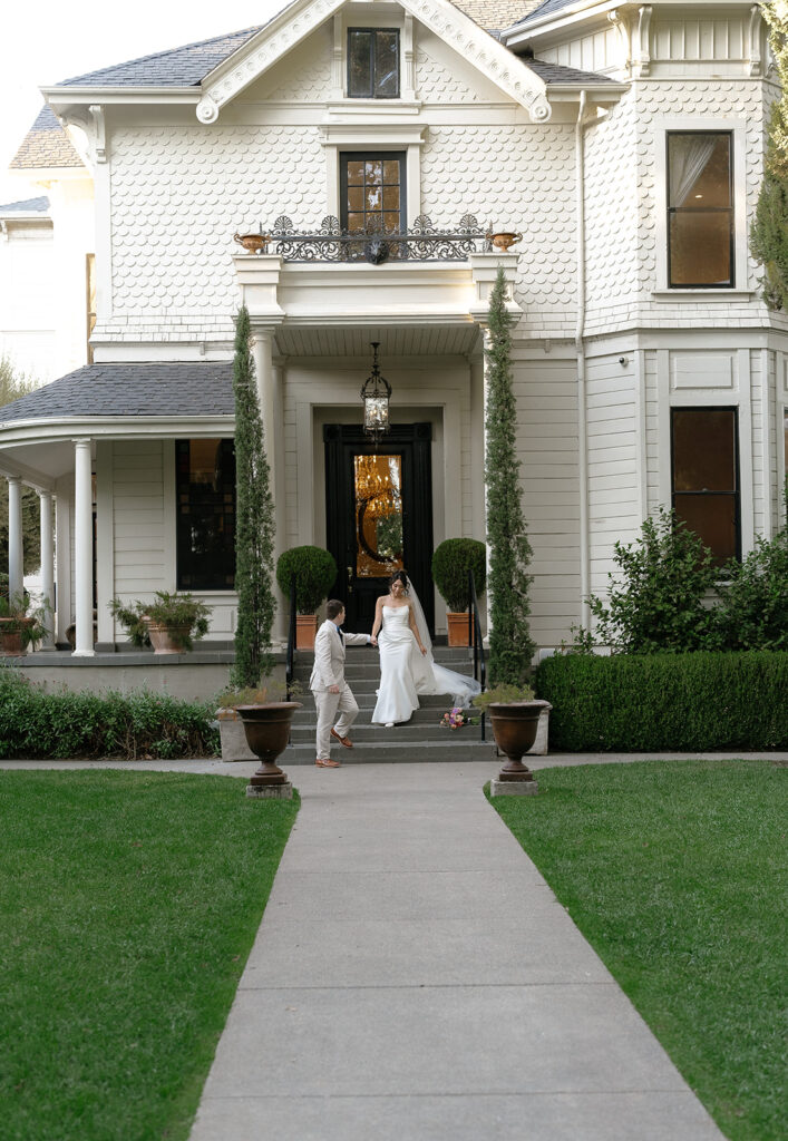 A bride and groom stand near the entrance of a large white Victorian house with manicured greenery and a sidewalk leading to the door.