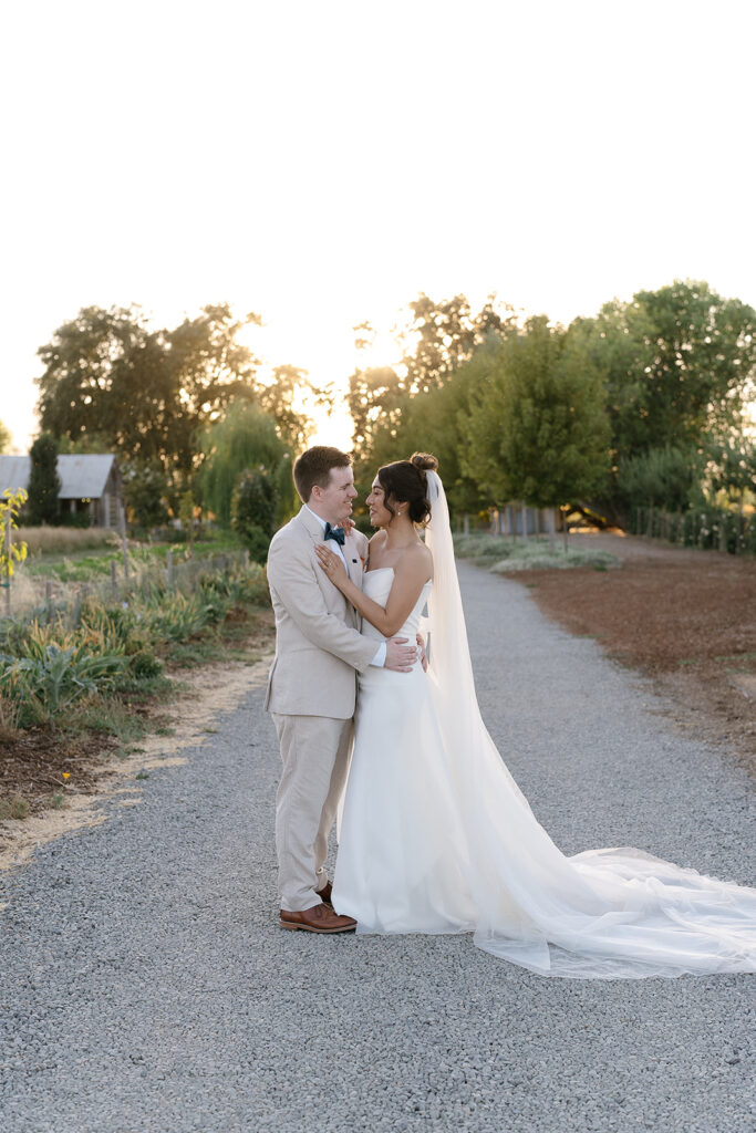 A bride and groom stand facing each other on a gravel path outdoors, embracing and smiling, with trees and sunlight in the background.