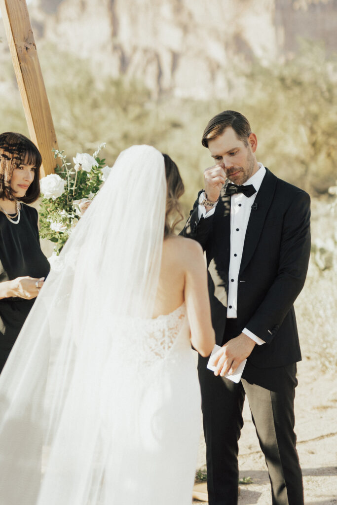 A groom in a black suit wipes a tear during a wedding ceremony outdoors. The bride, wearing a white dress and veil, stands in front of him. A woman holding a book is present beside them.