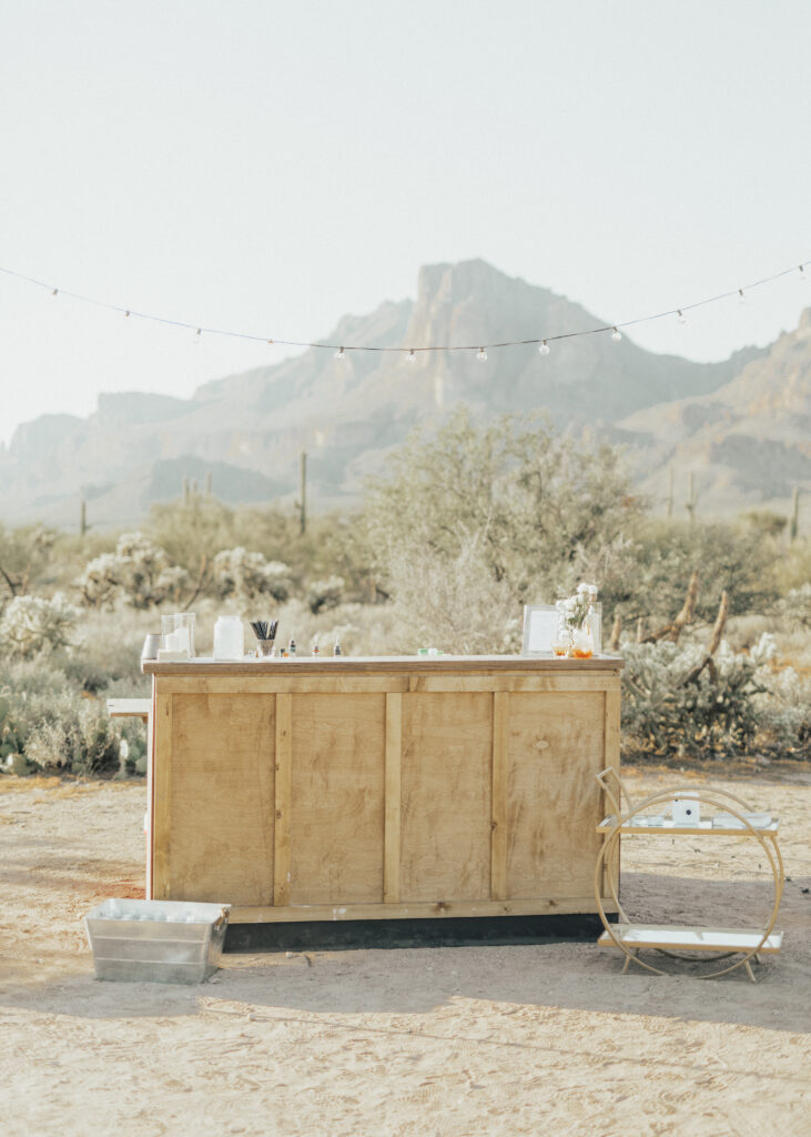 A wooden outdoor bar setup in a desert landscape, with mountains in the background and string lights overhead.