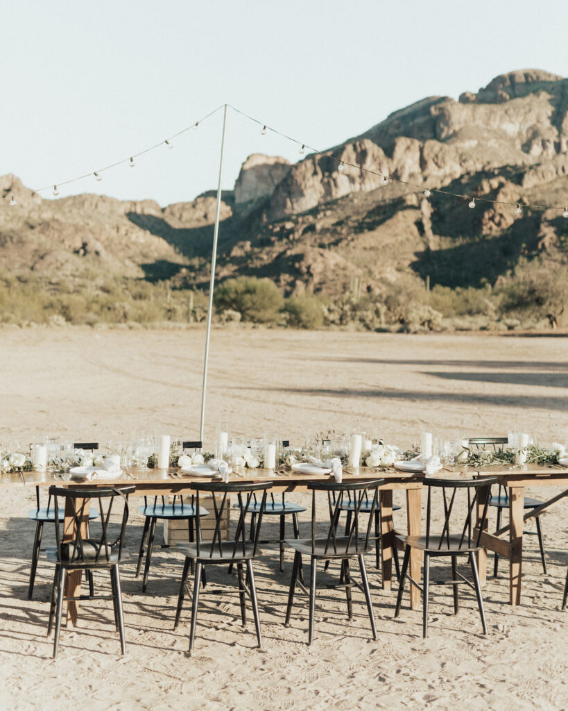 Outdoor dining setup on a sandy ground with wooden tables and chairs, decorated with candles and greenery. String lights hang above, with mountains visible in the background.